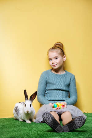 Cute smiling child girl sitting on a grass with Easter bunny and painted eggs. Easter holidays. Children's fashion. Happy yellow background.の写真素材