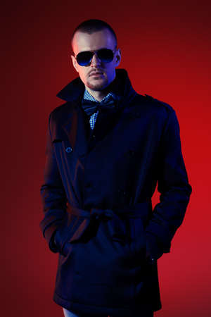Fashionable male model wearing coat and sunglasses posing at studio over red background. Men's beauty, seasonal collection.の写真素材