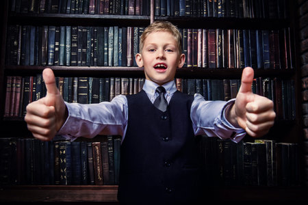 Smart boy in a suit stands in the library by the bookshelves and shows thumbs.