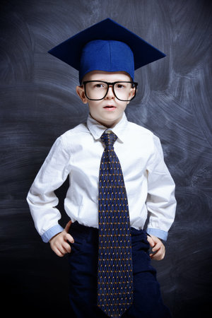 Five year old boy in white shirt, a tie, academic hat and big glasses standing by a blackboard....