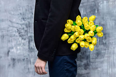 A man in elegant black suit hides a bouquet of tulips behind his back. Valentine's Day, Women's Day, Mother's Day. Wedding concept.の写真素材
