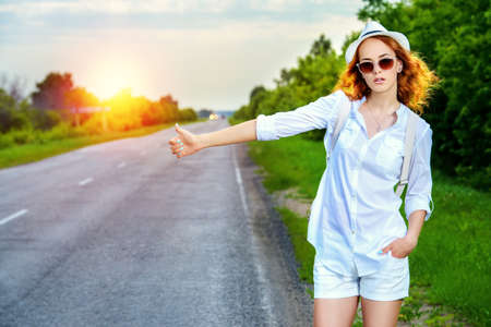Hitchhiking girl. Attractive young woman standing on a highway and catching a passing car.の写真素材