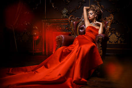 Magnificent young woman in luxurious red dress and precious jewelery is sitting in a chair in a luxury apartment. Classic vintage interior. Beauty, fashion.の写真素材