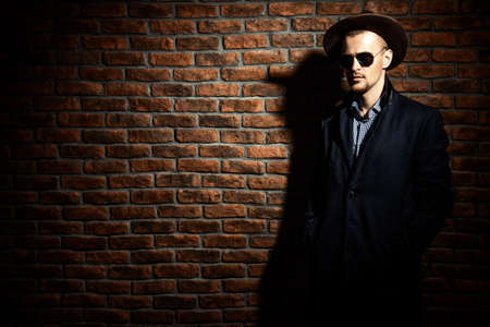 Portrait of a stylish handsome man wearing hat, coat and sunglasses. Men's beauty, seasonal fashion. Brick wall background.の写真素材
