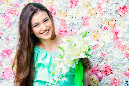 Beautiful smiling young woman in long light dress standing with a bouquet of white flowers by a floral background. The mood of the spring and summer. Beautiful smiling bride, wedding.の写真素材