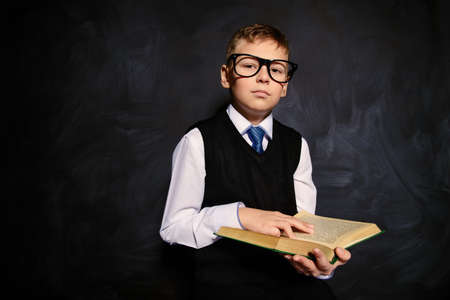 Smart schoolboy in elegant suit and spectacles standing with his book by a blackboard. Educational concept. Copy space.の写真素材