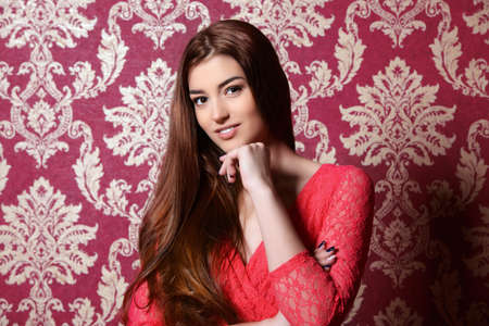 Beautiful young woman wearing evening red dress posing over vintage wallpaper. Beauty, fashion.の写真素材