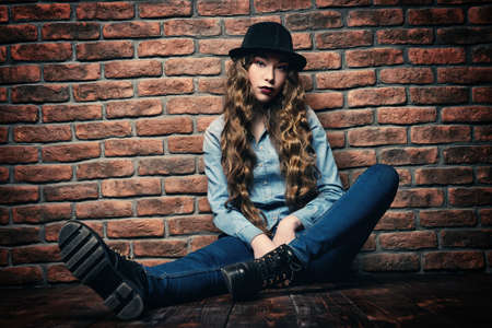 Attractive young woman sitting on a floor by the brick wall. Hipster girl. Youth fashion, beauty. Long hair, haircare.の写真素材