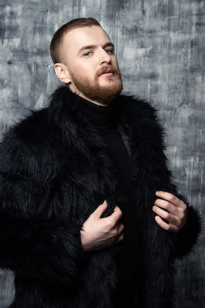 Fashion shot of a stylish brutal bearded man wearing fur coat. Studio shot over grunge background.