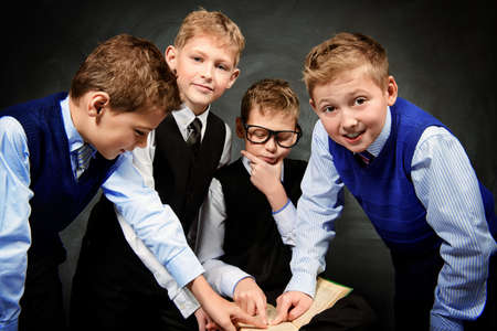 Group of smart boys in formal suits enthusiastically read the book by a blackboard. Educational concept. Copy space.の写真素材
