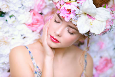 Beautiful romantic young woman in a wreath of flowers posing on a background of roses. Inspiration of spring and summer. Perfume, cosmetics concept.の写真素材