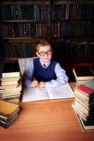 Smart boy in a suit sitting in the library by the bookshelves with many old books. Educational...