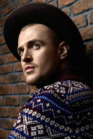 Portrait of a handsome man wearing sweater and hat standing by the brick wall. Male beauty, fashion.の写真素材