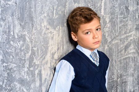 Portrait of a sad boy. Seven year old boy in elegant school clothes standing by a grey wall with a sad face. Loneliness.の写真素材