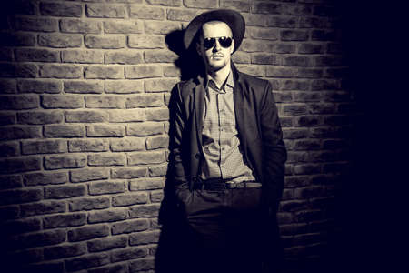 Portrait of a stylish handsome man wearing hat, coat and sunglasses. Men's beauty, seasonal fashion. Brick wall background.の写真素材
