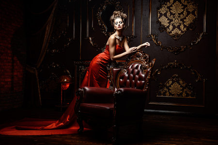 Magnificent young woman in luxurious red dress and precious jewelery posing in a luxury apartment. Classic vintage interior. Beauty, fashion.の写真素材