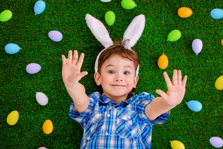 Funny little boy with bunny ears lying on a grass among colorful Easter eggs.の写真素材