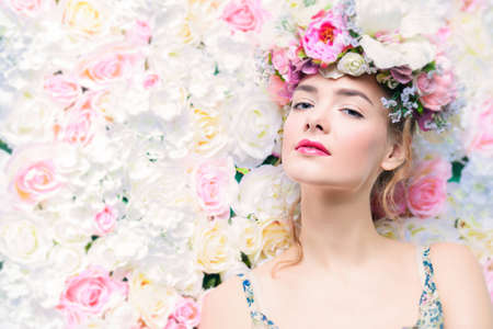 Beautiful romantic young woman in a wreath of flowers posing on a background of roses.の写真素材