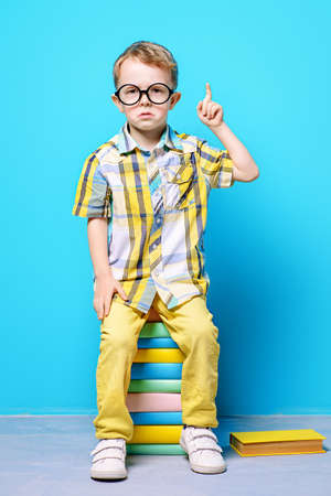 Cute serious little boy in bright clothes and big glasses sitting on a stack of books. Educational concept.の写真素材
