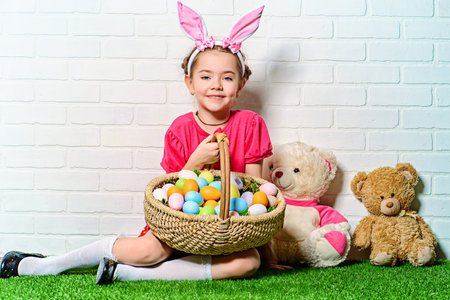 Cute happy little girl wearing bunny ears sits with Easter eggs on a green grass. Kid's fashion....