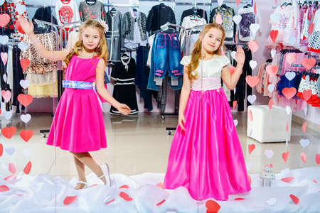 Two cute little girls in beautiful dresses posing in the window of children's clothing store. Kid's fashion. Seasonal sale and shopping.の写真素材