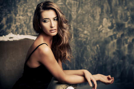 Fashion shot of a beautiful young woman in black dress posing on a vintage armchair. Femininity. Sexual woman. Beauty, fashion and cosmetics concept.の写真素材