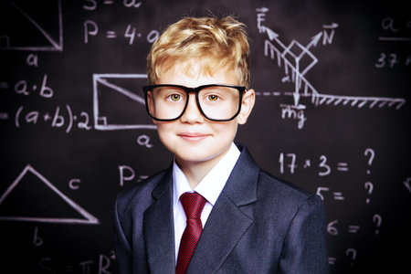 Funny smart boy in formal suit and glasses over school blackboard. Educational concept.の写真素材