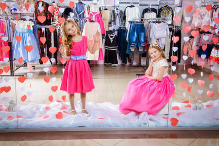 Two cute little girls in beautiful dresses posing in the window of children's clothing store. Kid's fashion. Seasonal sale and shopping.の写真素材