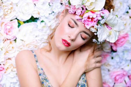 Beautiful romantic young woman in a wreath of flowers posing on a background of roses. Inspiration of spring and summer. Perfume, cosmetics concept.の写真素材