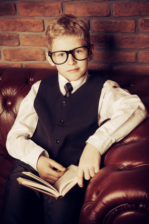 Smart boy in elegant suit and glasses sitting on a Chesterfield sofa with a book. Educational...