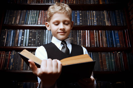 Smart boy in a suit sitting in the library by the bookshelves with many old books. Educational...