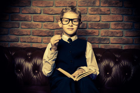Smart boy in elegant suit and glasses sitting on a Chesterfield sofa with a book. Educational...