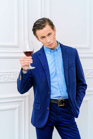 Good looking young man with a glass of red wine standing in luxurious apartments. Men's beauty, fashion model. Sommelier.の写真素材