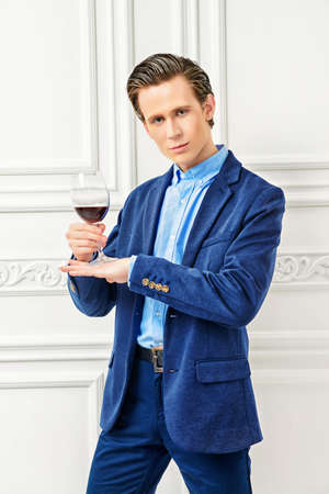 Good looking young man with a glass of red wine standing in luxurious apartments. Men's beauty, fashion model. Sommelier.の写真素材