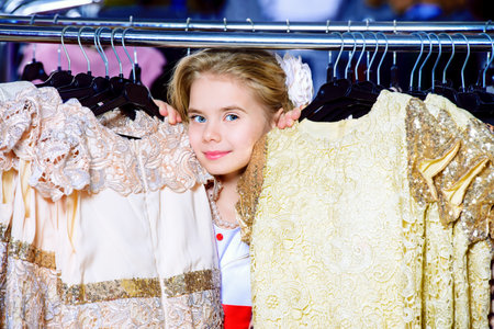 Cute little girl among dresses in a clothing store. Children's fashion. Sale and shopping concept.