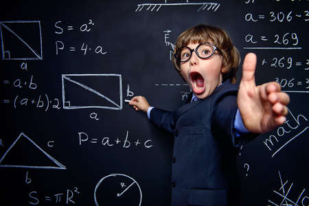 Smart schoolboy in black suit and glasses explaning something emotionally at the blackboard. Educational concept.の写真素材