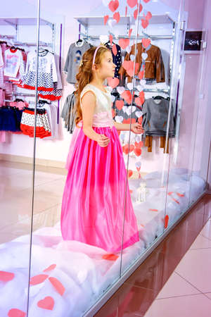 Cute little girl in a beautiful dress posing in the window of children's clothing store. Kid's fashion. Seasonal sale and shopping.の写真素材