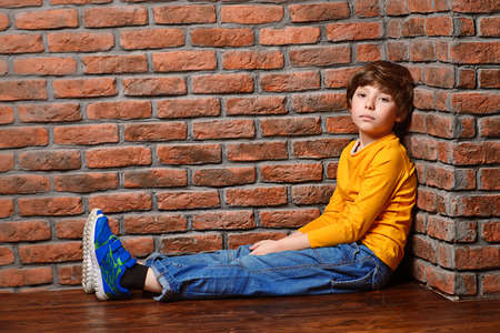 Portrait of a cute nine year old boy sitting on a floor leaning against the brick wall. Clothes for children. Kid's fashion.の写真素材