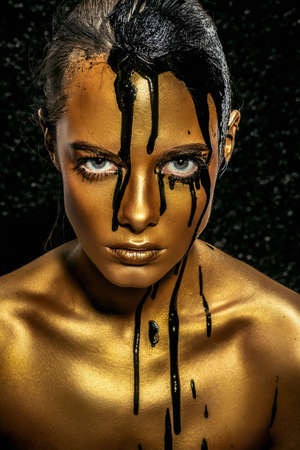 Beauty provocation. Body painting project. Portrait of a young woman with golden skin and black oil pouring on her. Fashionable provocation. Pollution and poisoning concept.の写真素材
