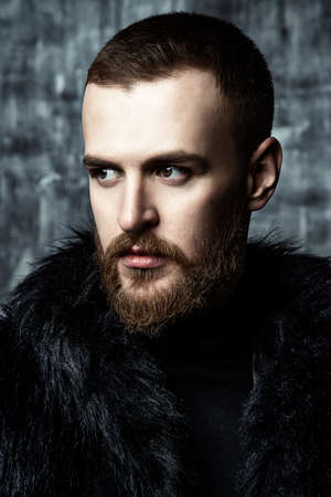 Fashion shot of a stylish brutal bearded man wearing fur coat. Studio shot over grunge background.の写真素材