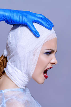 Beauty, fashion and medicine, plastic surgery. Portrait of a shouting young woman in bandages and hospital gown. Studio shot.の写真素材