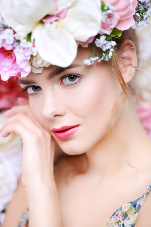 Beautiful romantic young woman in a wreath of flowers posing on a background of roses.の写真素材