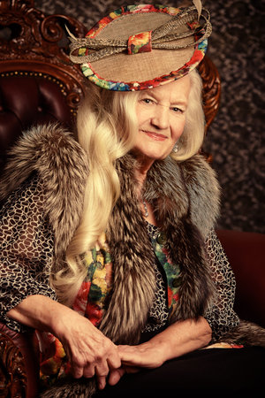 Fashionable old woman with beautiful blonde hair and elegant hat posing in vintage interior.