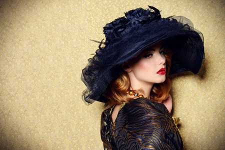 Portrait of a gorgeous young woman in elegant broad-brimmed hat and luxurious dress over golden background. Vintage style. Beauty, fashion.の写真素材