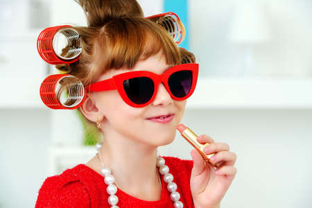 Portrait of a funny little girl with curlers in her hair paints lips with lipstick. Kid's fashion. Family at home.の写真素材
