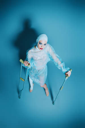 Fashion shot. Gorgeous female model in bandages and hospital gown posing at studio with crutches. Beauty and medicine, plastic surgery.の写真素材
