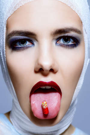 Close-up portrait of an attractive young woman in bandages holding a pill capsule in her mouth. Beauty, fashion and medicine, plastic surgery. Studio shot.の写真素材