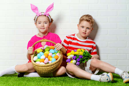 Cute happy children wearing bunny ears sits with Easter eggs and flowers on a green grass. Kid's fashion. Easter holidays.の写真素材