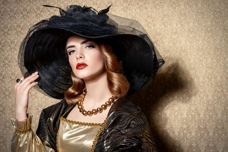 Portrait of a gorgeous young woman in elegant broad-brimmed hat and luxurious dress over golden background. Vintage style. Beauty, fashion.の写真素材