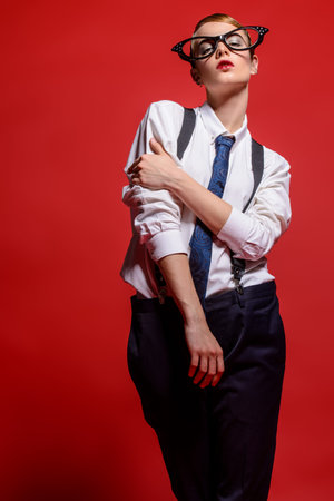Fashion shot. Attractive female model posing in a man's shirt and trousers with suspenders and elegant feminine glasses. Man's and women's style clothing. Red background.の写真素材